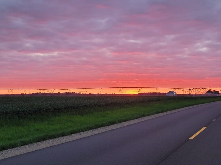 Sunset over agricultural fields with irrigation equipment and farm buildings along a rural road.