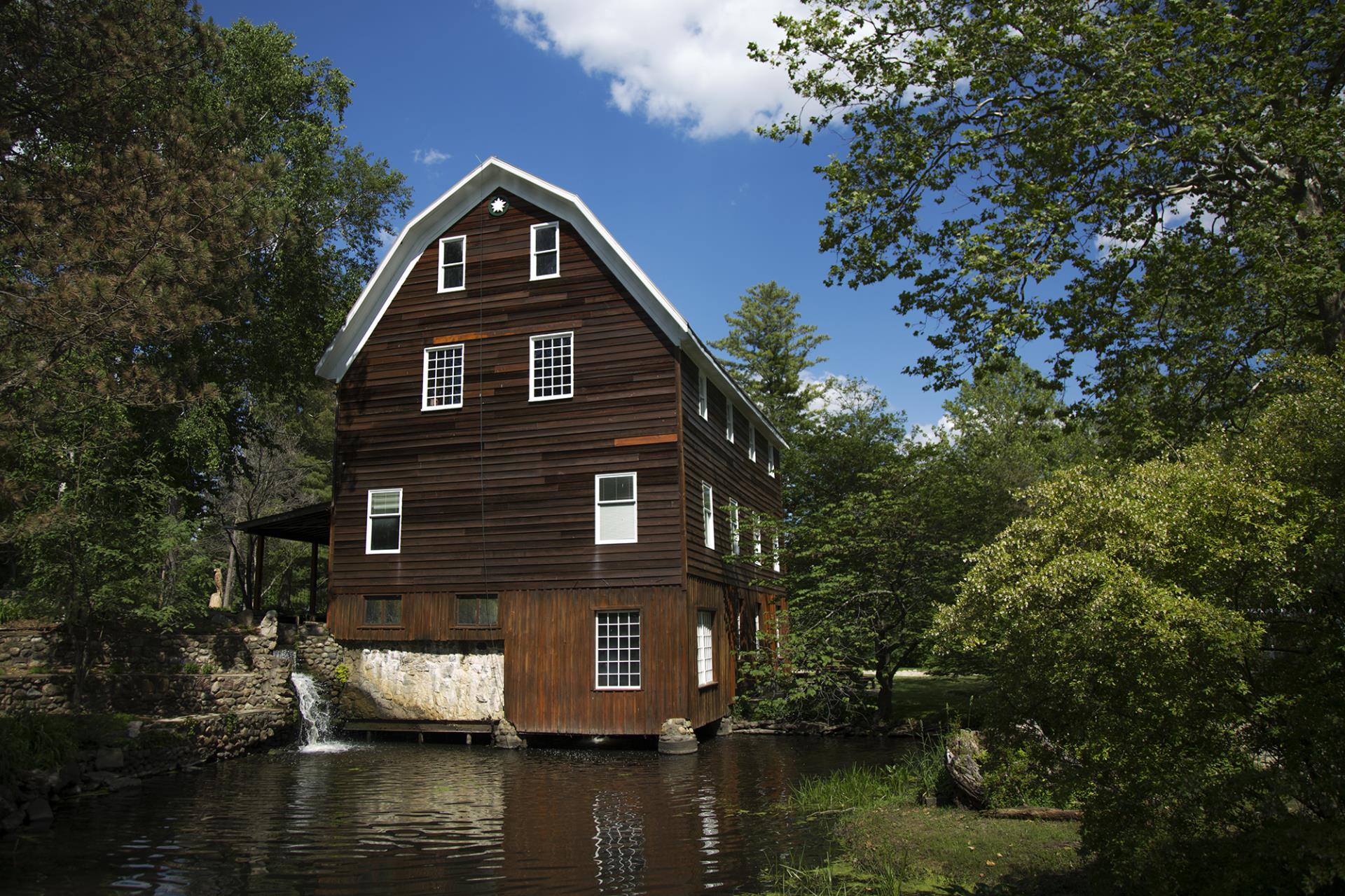 Brown wooden mill house with white trim situated over a stream, surrounded by green trees under blue sky.