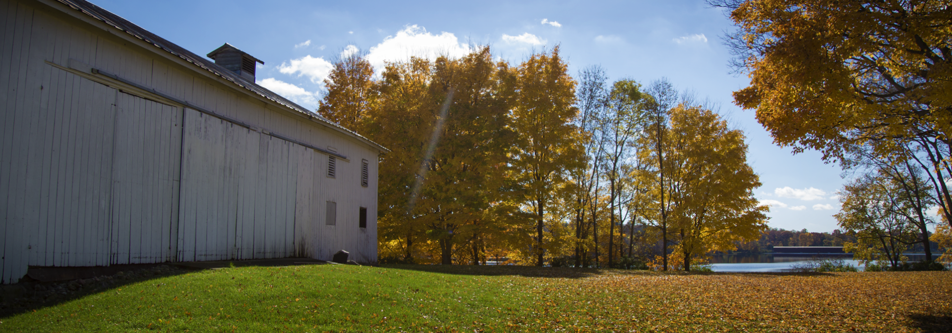 Historic white barn with cupola beside grassy field, autumn trees, and distant waterway under blue sky.
