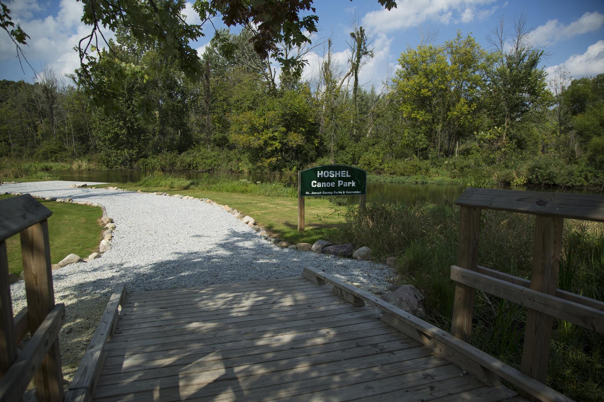 Hoshel Canoe Park entrance with wooden boardwalk, gravel path, and river surrounded by dense forest.