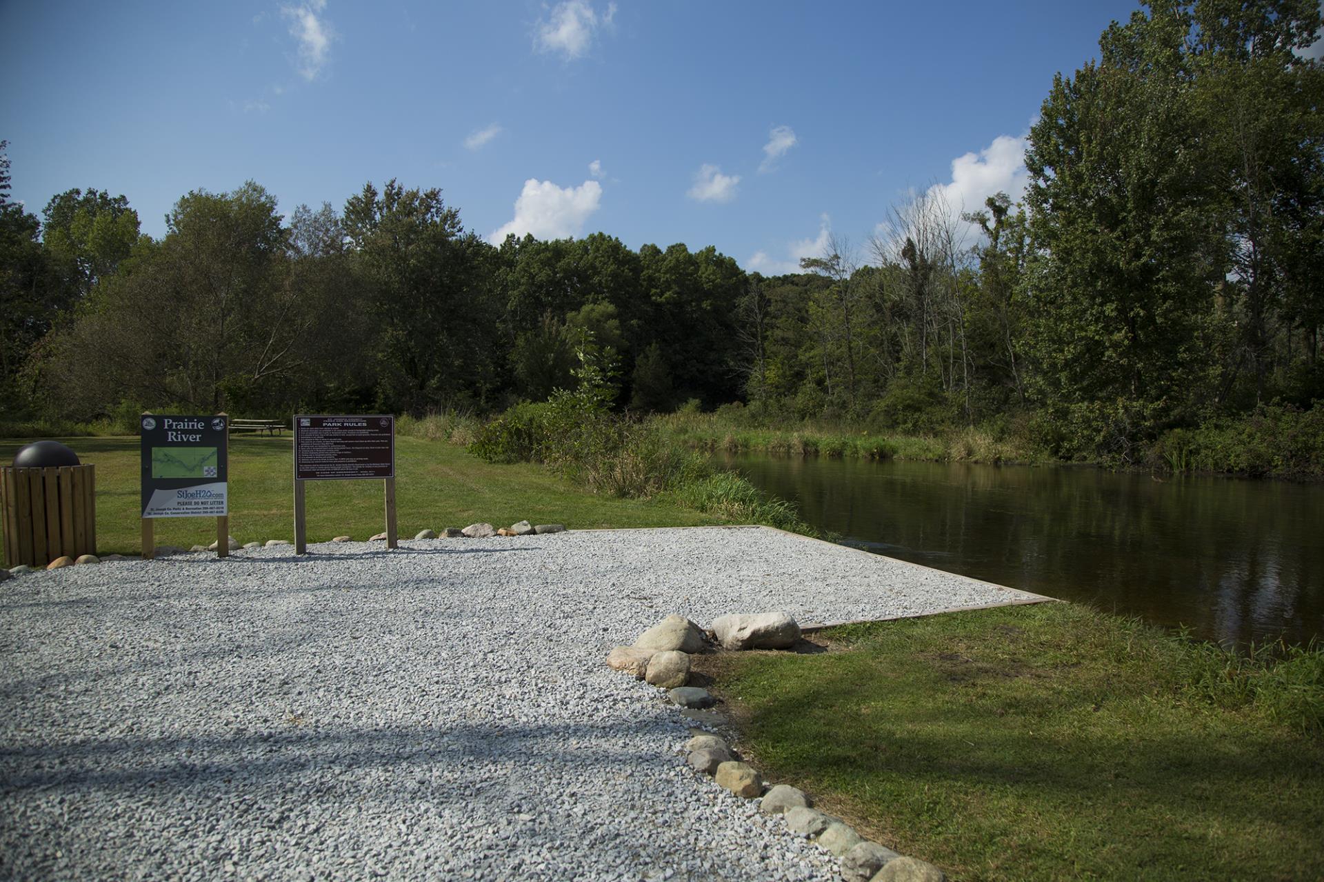 Prairie River overlook with informational signs, gravel area, and river view bordered by trees under clear sky.