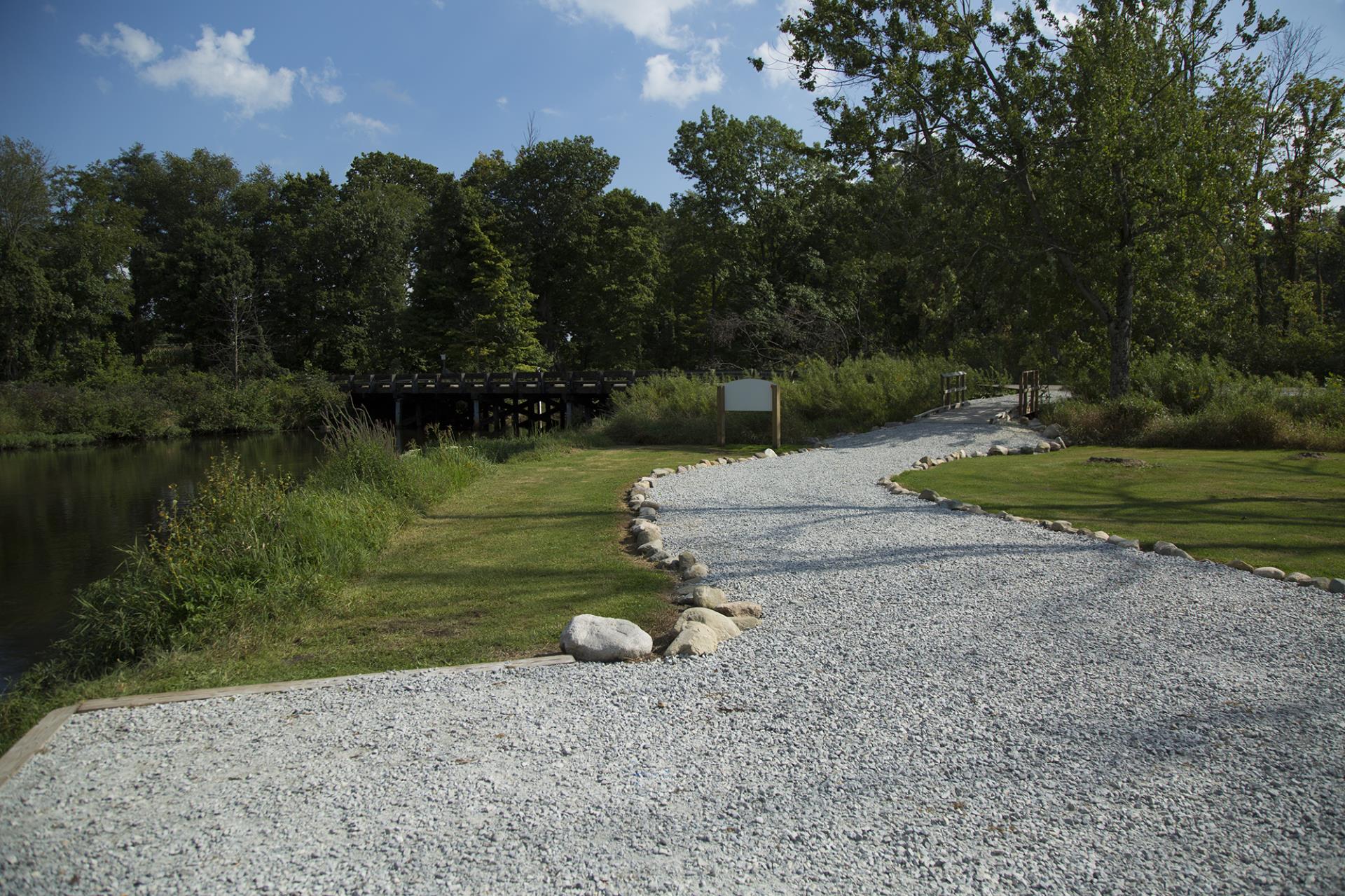 A winding gravel pathway along a waterfront with green lawn, trees, and wooden structures in the background under blue sky.