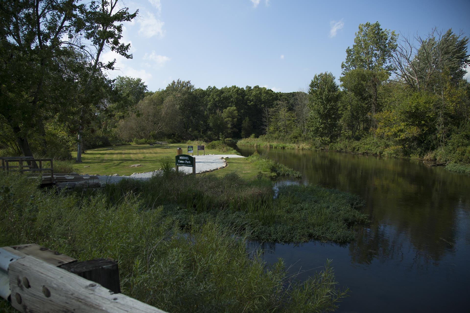 Hoshell Canoe Park entrance with river, grassy area, wooden dock, and surrounding trees under blue sky.