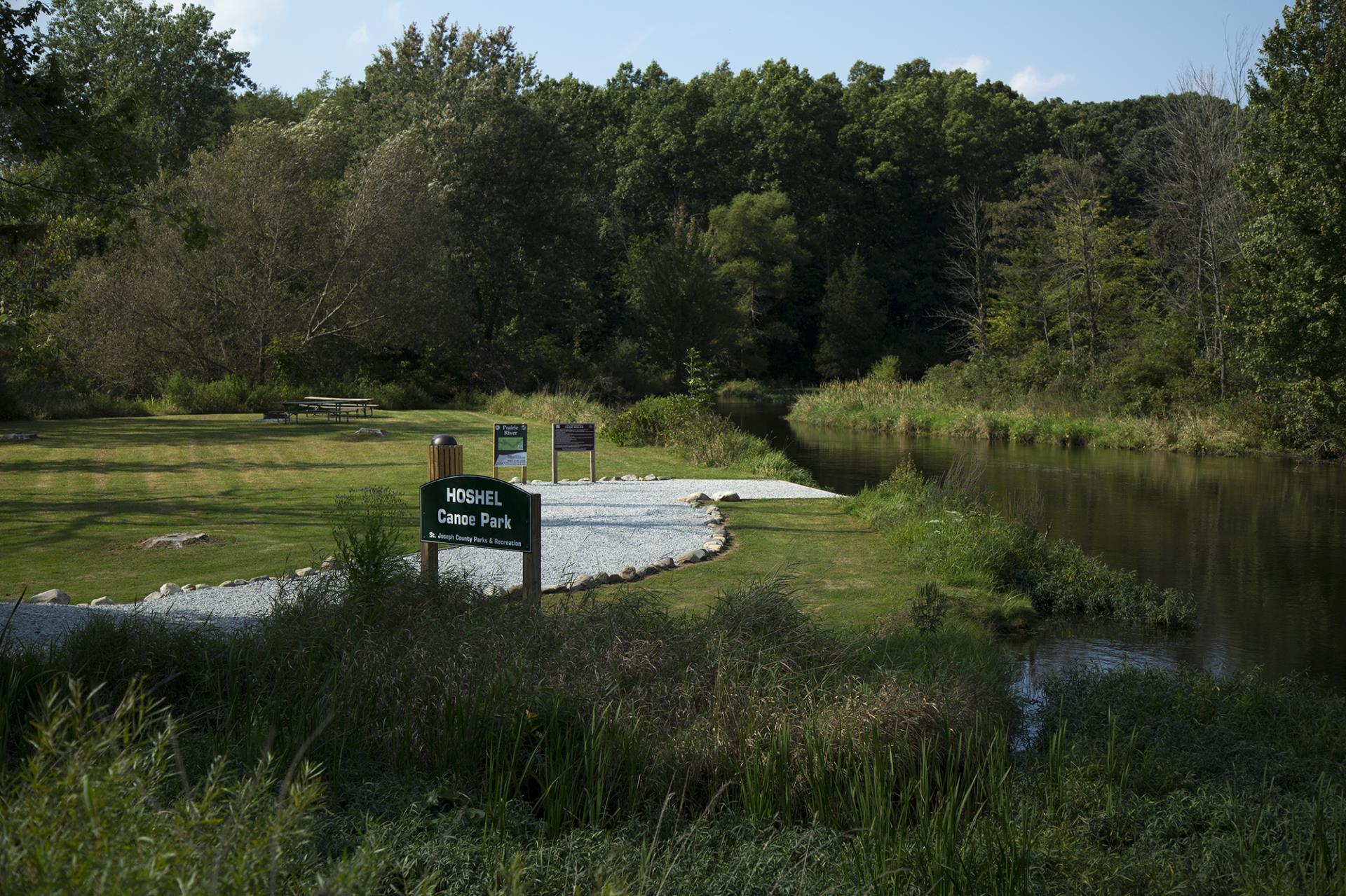 Hoshel Canoes Park entrance sign beside a river with gravel landing area, picnic tables, and forested shoreline.