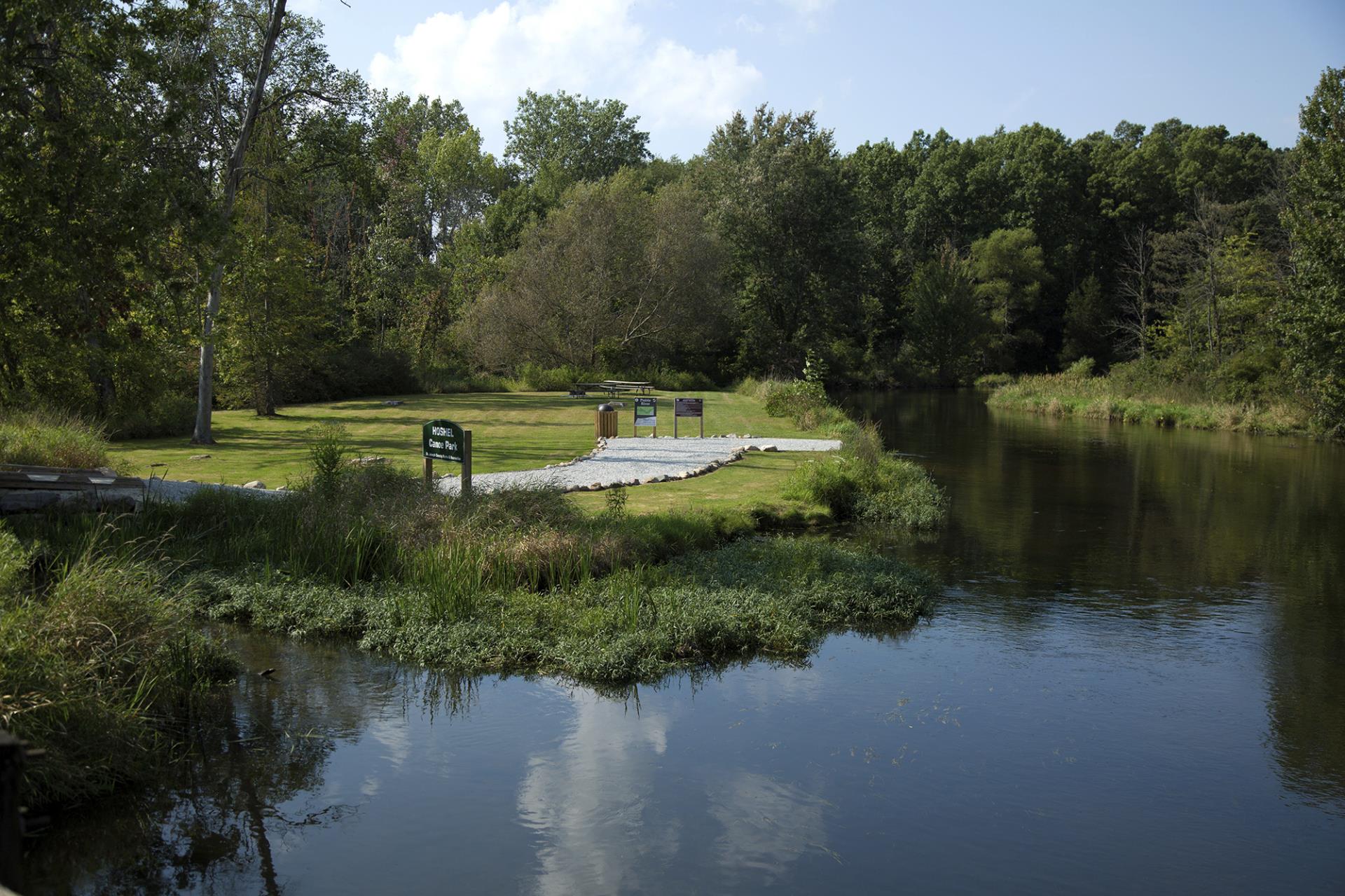 Riverside park with wooden dock, green lawn, interpretive signs, and dense forest under blue sky with clouds.
