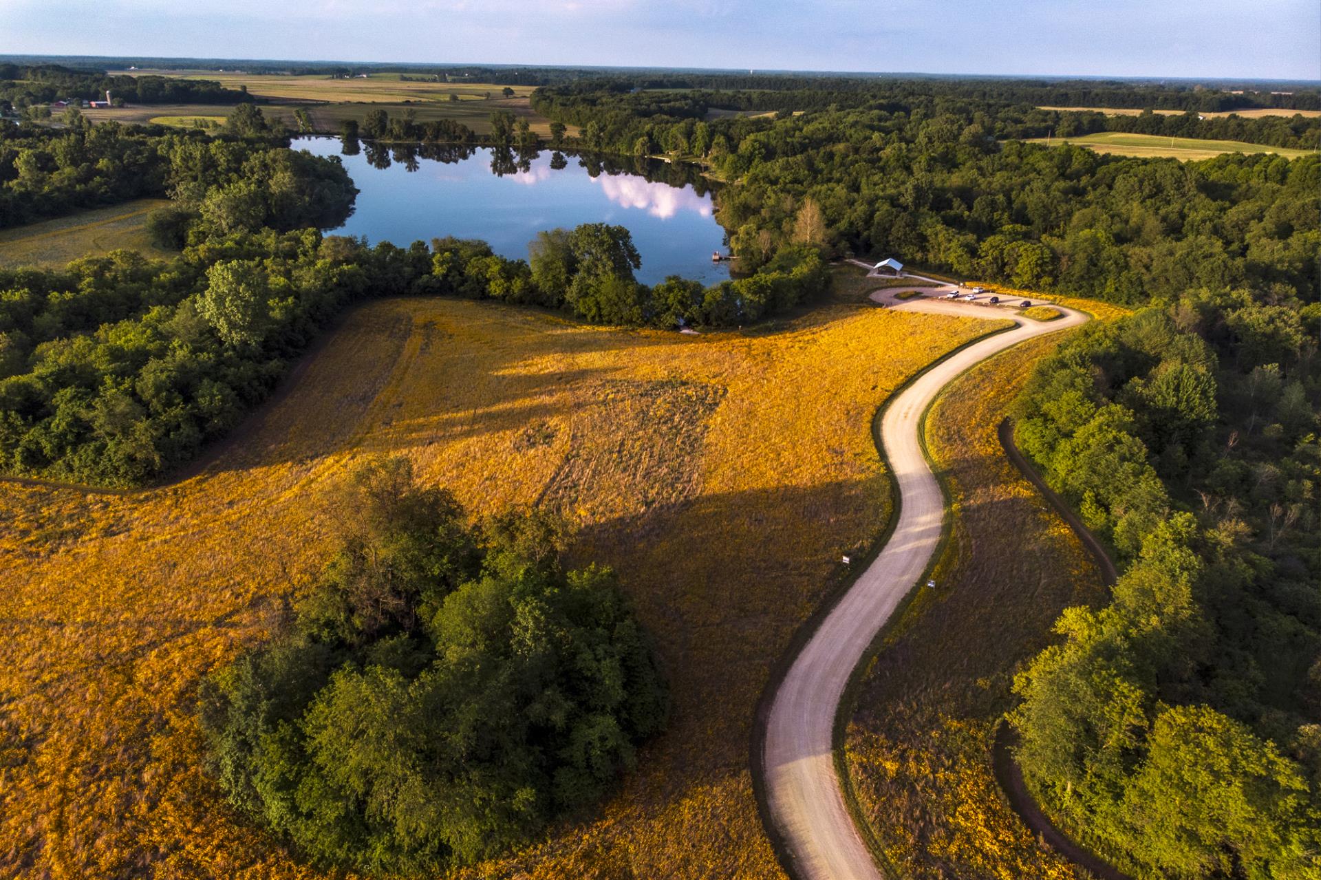 Aerial view of a winding rural road through farmland with a lake surrounded by trees in the distance.