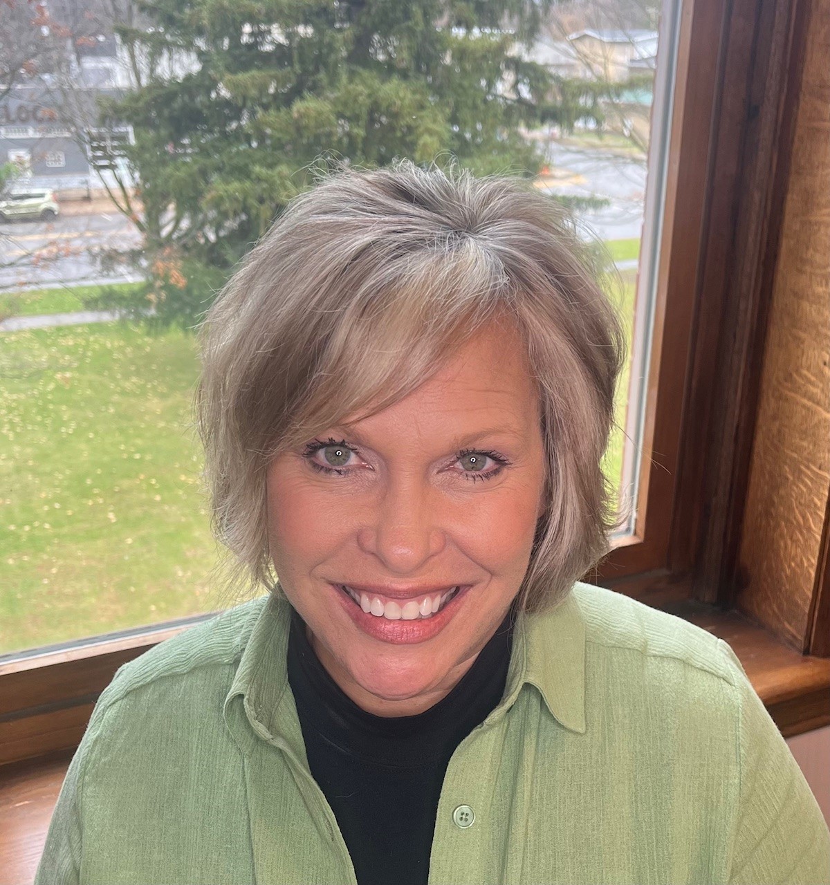 Woman with gray hair and green shirt smiling at camera, window with lawn and trees visible in background.